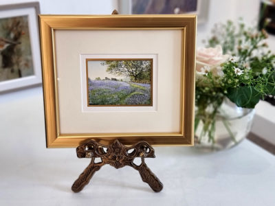 Rosalind Pierson art at Norton Way Gallery Hertfordshire. This beautiful, miniature, painting has been painted in watercolour. It is an original artwork from Rosalind Pierson and depicts a field of Bluebells and trees. The painting is shown here, on a miniature easel.