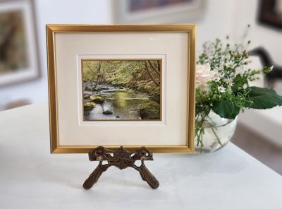 Rosalind Pierson art at Norton Way Gallery Hertfordshire. This beautiful, miniature, painting has been painted in watercolour. It is an original artwork from Rosalind Pierson and depicts a shaded summer river. There are trees and bolders. This miniature, watercolour is shown on a miniature easel.