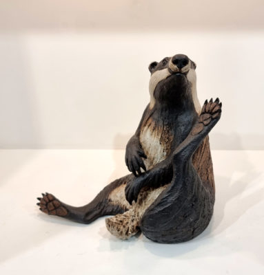 Pippa Hill at Norton Way Gallery Hertfordshire. This ceramic clay badger is an original artwork from Pippa Hill. The badger is sitting up with legs a kimbo and he is scratching his chin, he is white, black and brown.