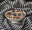 Anne Songhurst Art at Norton Way Gallery Hertfordshire. This beautiful oil painting is an original artwork by British artist Anne Songhurst. It is a still life painting, depicting a white bowl of Liquorice Allsorts on black and white, striped material.